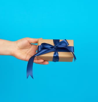 hand holds a box with a gift wrapped in brown kraft paper and tied with a silk blue ribbon on a blue background, concept of giving a gift
