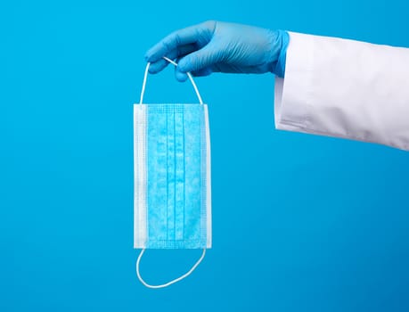 doctor in blue latex sterile gloves holds a blue disposable mask made of non-woven material on a blue background, protective accessory for the respiratory tract from the virus