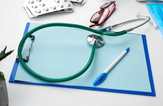 metal stethoscope, paper holder with a blank blue sheet, disposable blue medical mask on a white background, top view