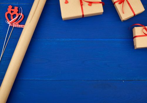 Wrapped gifts in brown craft paper and tied with a red ribbon, a roll of paper and scissors on a blue wooden background, place for text in the middle