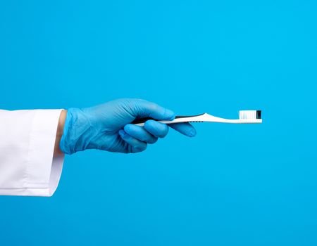 doctor therapist is dressed in a white robe uniform and blue sterile gloves holding a toothbrush, daily brushing concept, blue background