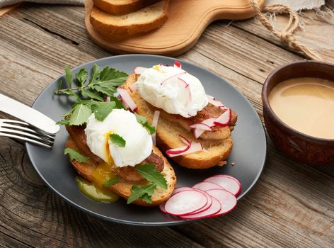 two sandwiches on a toasted white slice of bread with poached eggs, green leaves of arugula and radish in a round plate, behind a cup of coffee, morning breakfast