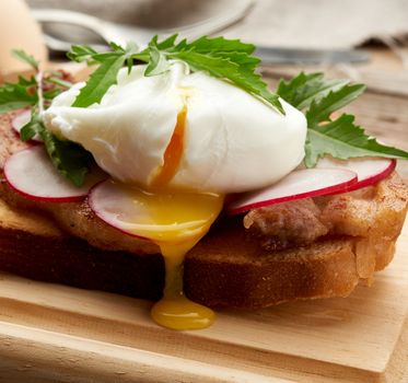 sandwich on toasted white slice of bread with poached eggs, green leaves of arugula and radish, morning breakfast on a brown wooden board