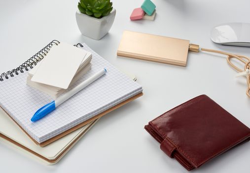 notebooks, pen, stack of business cards, brown leather wallet and power bank with a cord on a white table, workplace, top view