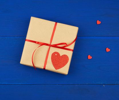 gift wrapped in brown kraft paper and tied with a thin red ribbon on a blue wooden background, top view, flat lay. Eco package design