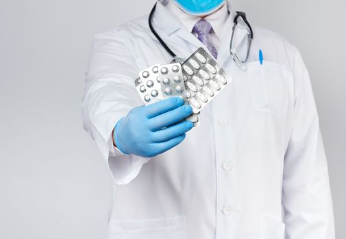 adult doctor therapist is dressed in a white uniform coat and blue sterile gloves is standing and holding a stack of pills in blister packs, white background