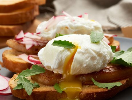 sandwich on toasted white slice of bread with poached eggs, green leaves of arugula and radish, morning breakfast on a brown wooden board, close up