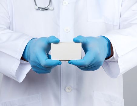 adult doctor therapist is dressed in a white robe uniform and blue sterile gloves is standing and holding a stack of empty white paper business cards, place for inscription