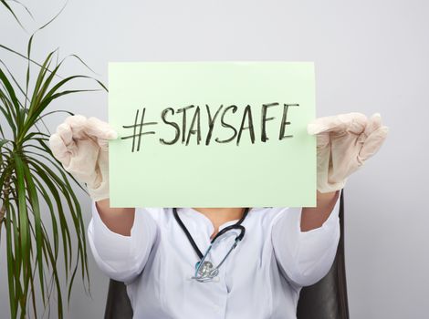young woman doctor in a white coat, sterile medical gloves holds a poster with the inscription stay safe, concept of self-isolation during a pandemic and epidemic