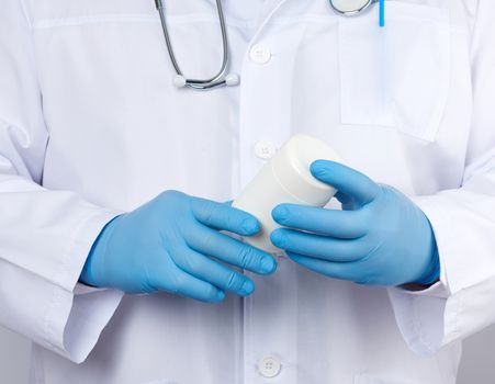 male doctor in a white medical coat and blue latex gloves holds a white plastic jar of pills, close up