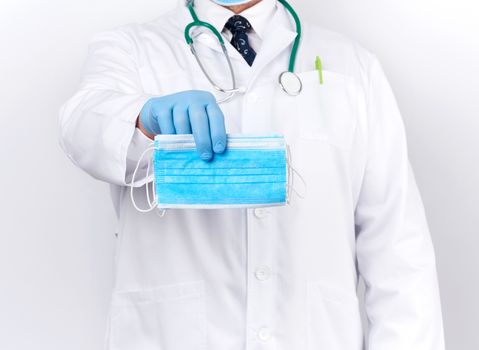 doctor in a white coat, blue latex sterile gloves holds textile medical masks in his hand on a white background, protective accessory against viruses and bacteria