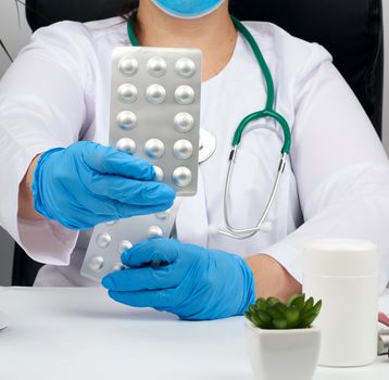 female hand with blue sterile gloves holds a stack of pills in a blister, concept for drug treatment of diseases, doctor sits in an armchair at a desk, office