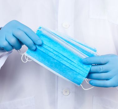doctor in a white coat, blue latex sterile gloves holds textile medical masks in his hand, protective accessory against viruses and bacteria, close up