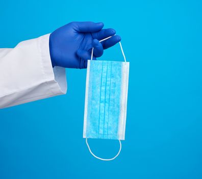 doctor in a white coat, blue latex sterile gloves holds textile disposable medical masks in his hand on a blue background, additional protection against viruses during an epidemic, pandemic
