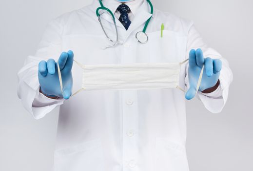 doctor in a white coat, blue latex sterile gloves holds textile medical masks in his hand on a white background, homemade mask made of white gauze and elastic