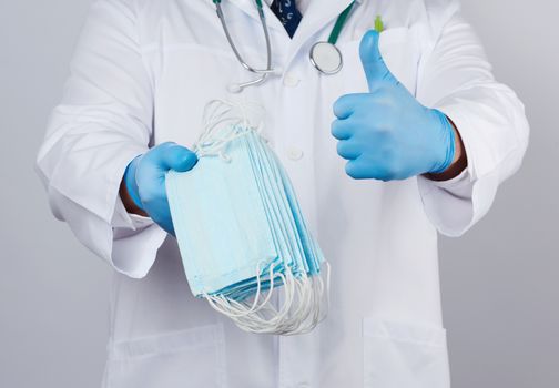doctor in a white coat, blue latex sterile gloves holds textile medical masks in his hand on a white background, other hand shows like, protective accessory against viruses and bacteria