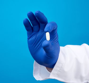 male hand in blue sterile gloves holds an oval white pill, medical treatment concept for diseases, blue background
