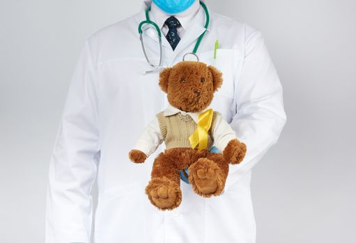 pediatrician in white coat, blue latex gloves holds a brown teddy bear with a yellow ribbon on a sweater, concept of the fight against childhood cancer, problem of suicides