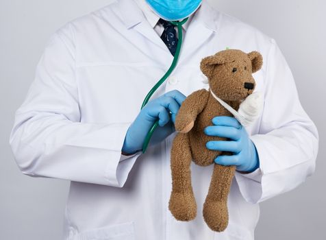 pediatrician in a white coat, blue latex gloves holds a brown teddy bear with a bandaged paw, doctor examines the toy with a stethoscope, white background