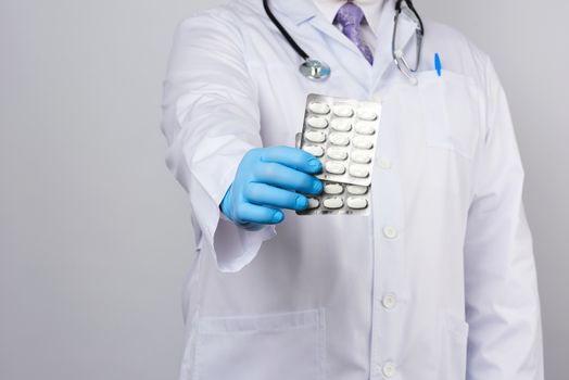 adult doctor therapist is dressed in a white uniform coat and blue sterile gloves is standing and holding a stack of pills in blister packs, white background