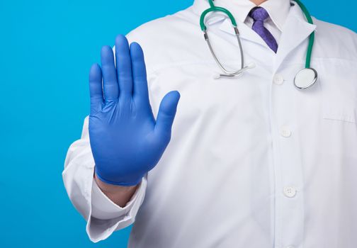 male doctor in a white coat and blue sterile gloves shows a stop gesture with his right hand, blue background
