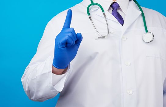 male doctor in white coat, blue medical gloves shows hand gesture of attention, index finger up