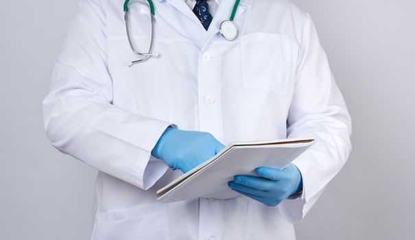 male doctor in a white coat with buttons, wearing blue sterile gloves, holding a paper notebook in his hand on a white studio background