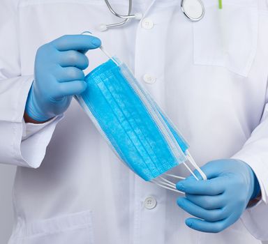 doctor in a white coat, blue latex sterile gloves holds textile medical masks in his hand, protective accessory against viruses and bacteria, close up