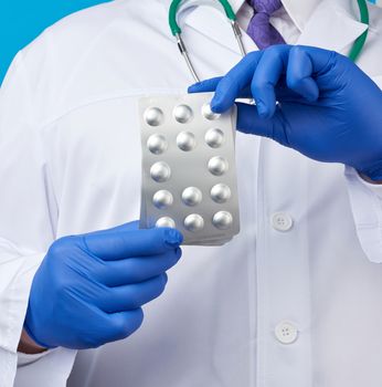 male hand with blue sterile gloves holds a stack of pills in a blister, concept for drug treatment of diseases, blue background