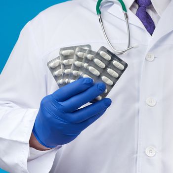 male hand with blue sterile gloves holds a stack of pills in a blister, concept for drug treatment of diseases, blue background