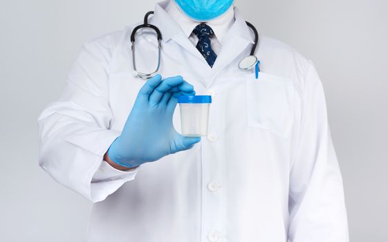 male doctor in a white coat and tie stands and holds a plastic container for urine specimen on a white background, wearing blue sterile medical gloves
