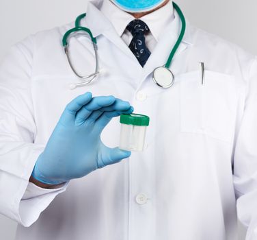 doctor in a white coat and blue sterile latex gloves holds a plastic jar for stool analysis, white background