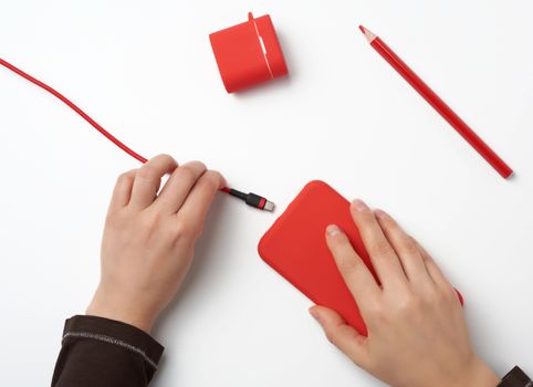 two hands holding a red mobile smartphone and a charging cable on a white background, top view