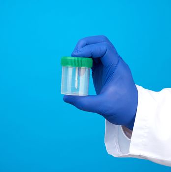 doctor in a white coat and blue sterile latex gloves holds a plastic jar for stool analysis, blue background