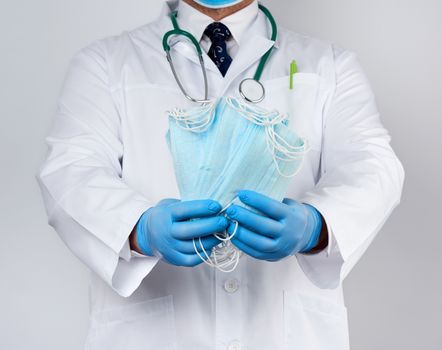 doctor in a white coat, blue latex sterile gloves holds textile medical masks in his hand on a white background, protective accessory against viruses and bacteria