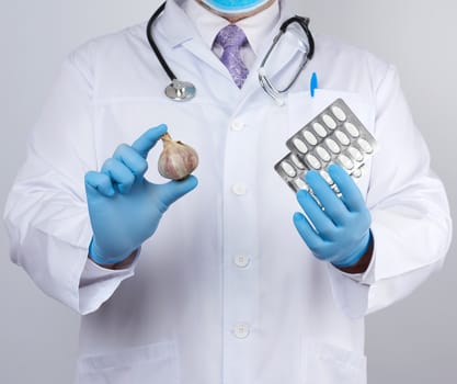 doctor in a white medical coat and blue rubber gloves holds a fresh head of garlic and pills, prevention against viral diseases, traditional medicine