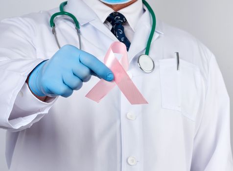 male doctor in a white coat and tie stands and holds a pink silk ribbon in the form of a loop, wearing blue sterile medical gloves, symbol of the fight against breast cancer in October 