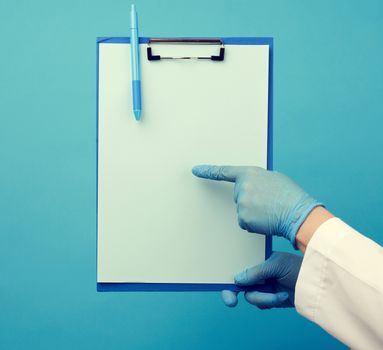 female doctor in a white coat, rubber blue medical gloves holds a folder for papers, blue background, copy space