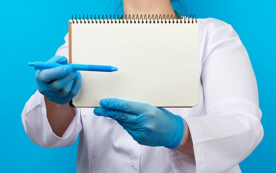 medic woman in white coat, blue latex gloves holding an open notebook with blank white sheets, blue background, place for text