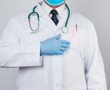 male doctor in a white coat and tie stands and holds a pink silk ribbon in the form of a loop, wearing blue sterile medical gloves, symbol of the fight against breast cancer in October 