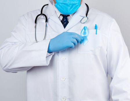 male doctor in a white coat and tie stands and holds a blue silk ribbon in a loop, wearing blue sterile medical gloves, symbol of the fight and treatment of prostate cancer