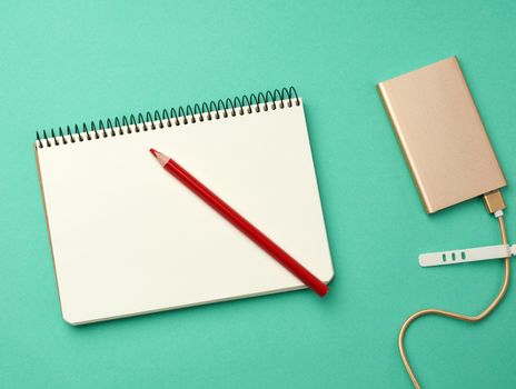 open notebook with blank white sheets and golden power bank and cord with a usb connector for recharging mobile devices on a green background, top view