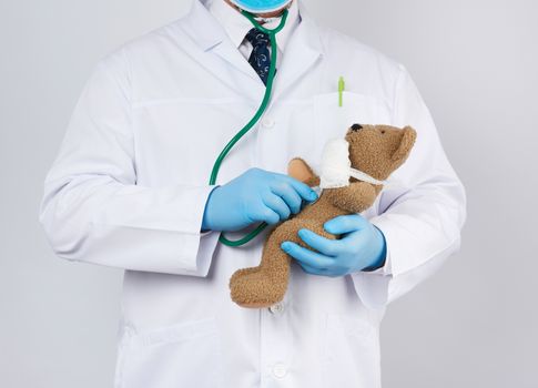 pediatrician in a white coat, blue latex gloves holds a brown teddy bear with a bandaged paw, doctor examines the toy with a stethoscope, white background