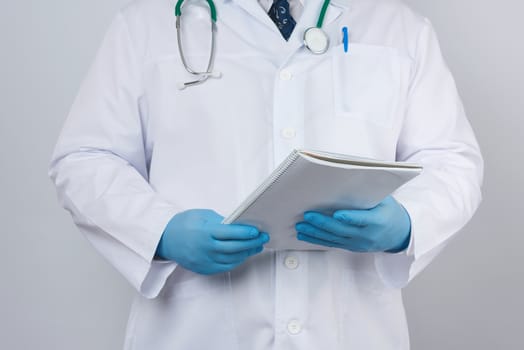 male doctor in a white coat with buttons, wearing blue sterile gloves, holding a paper notebook in his hand on a white studio background