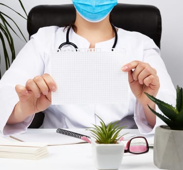 doctor in a white coat and medical blue gloves holds a blank white sheet, place for an inscription, workplace 