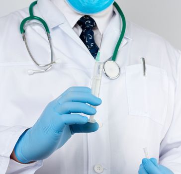 male doctor in a white coat and tie stands and holds a plastic syringe with a needle on a white background, wearing blue sterile medical gloves in his hands the concept of vaccination against diseases
