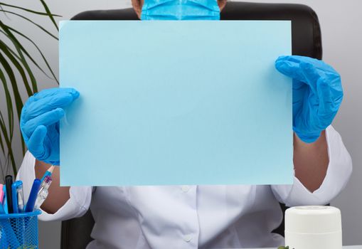 doctor in a white medical coat is sitting at a table in a brown leather chair and holding a empty blue sheet of paper in his hands, place for an inscription, medical office