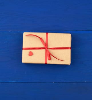 gift wrapped in brown kraft paper and tied with a thin red ribbon on a blue wooden background, top view, flat lay. Eco package design