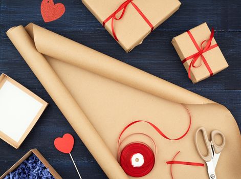 roll of brown kraft packaging paper, box tied with a red silk ribbon, scissors and a bobbin with ribbon on a blue wooden background, top view