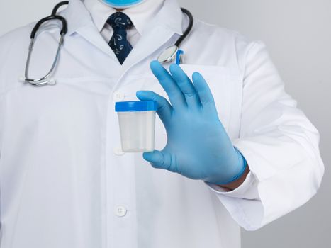 male doctor in a white coat and tie stands and holds a plastic container for urine specimen, wearing blue sterile medical gloves